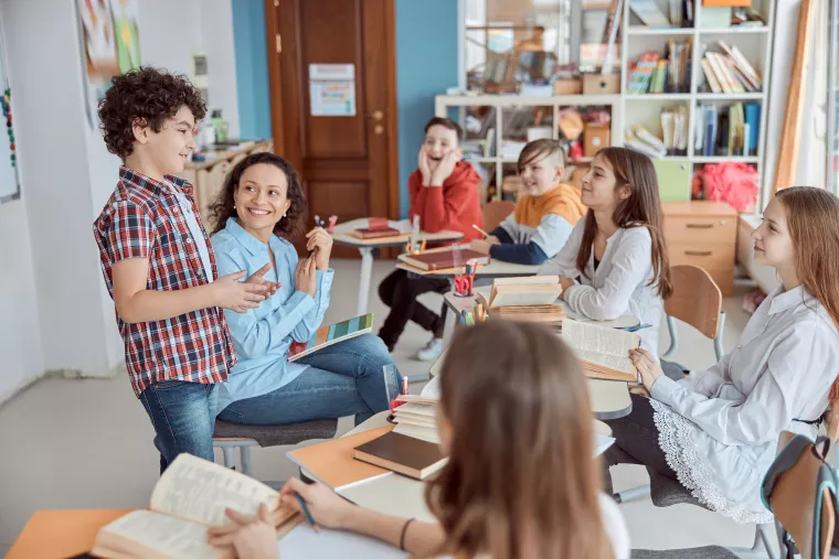 A young student talks in front of his classmates while standing next to his seated teacher.